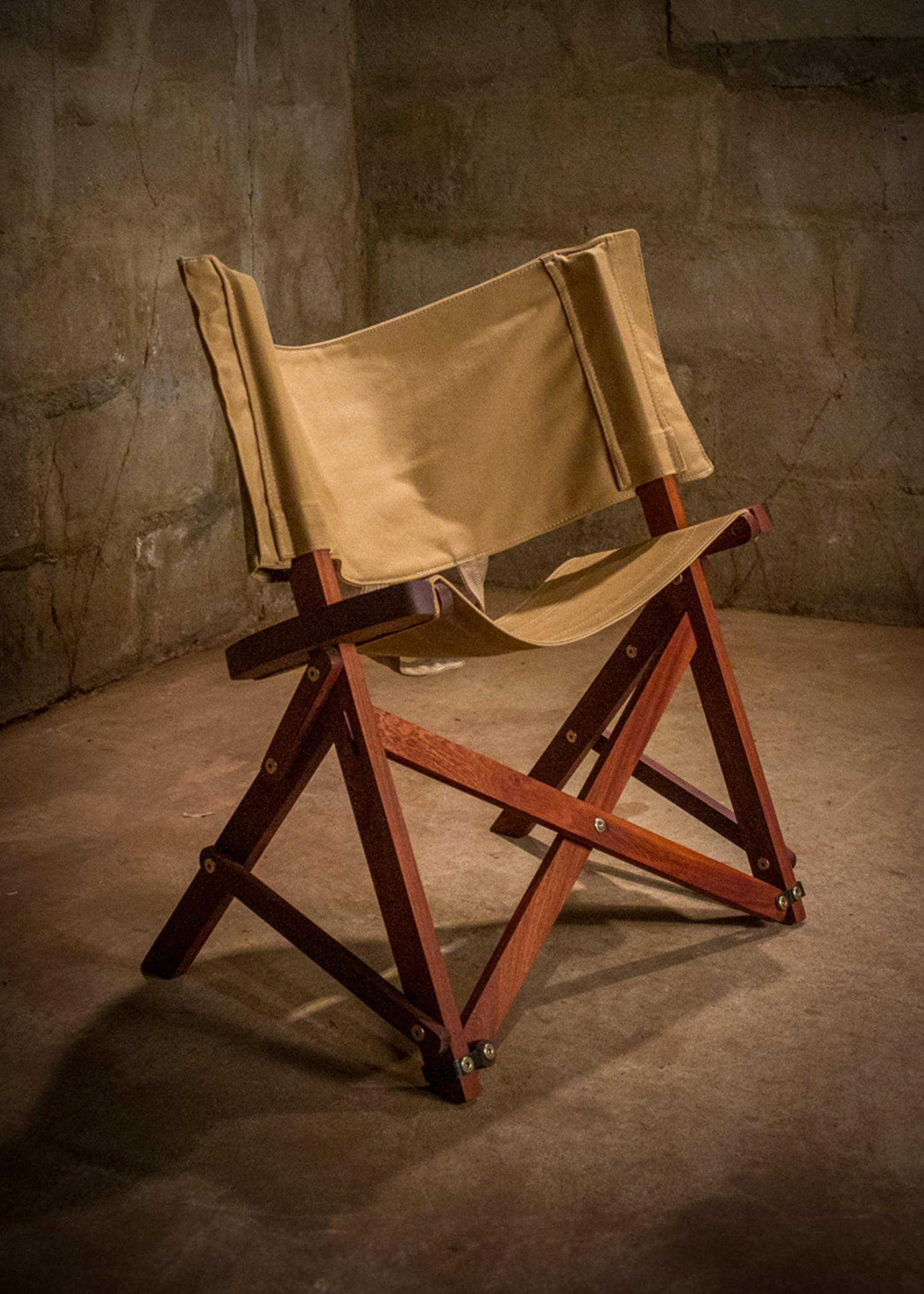 Classic Safari chair crafted with a rich mahogany wood frame and a khaki canvas seat and backrest, set in an ambient room with a textured concrete wall, exuding timeless style and handcrafted quality.