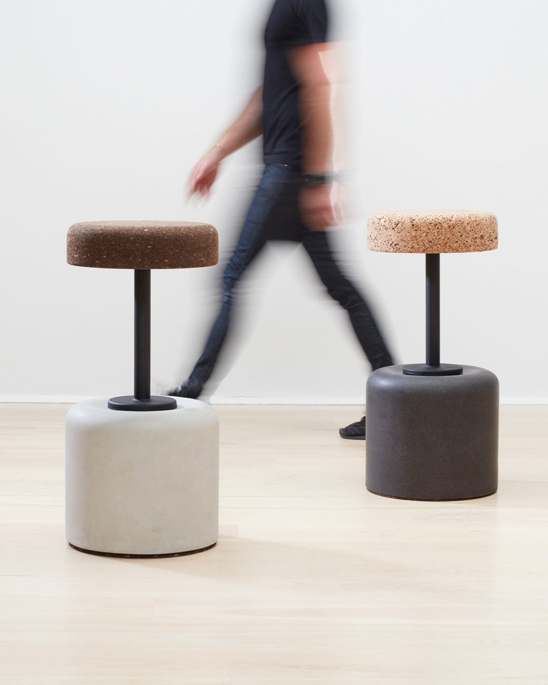 Designer Laurie Wiid van Heerden passes behind the elegantly designed Wiid Swivel Cork and Concrete Barstool in Natural Grey and Dark Cork, providing a sense of scale. This image emphasizes the stool's sleek, modern design and the contrast between the robust grey concrete base and the warm, dark cork seat. Laurie's presence highlights the thoughtful craftsmanship and sustainable innovation behind this contemporary piece of furniture