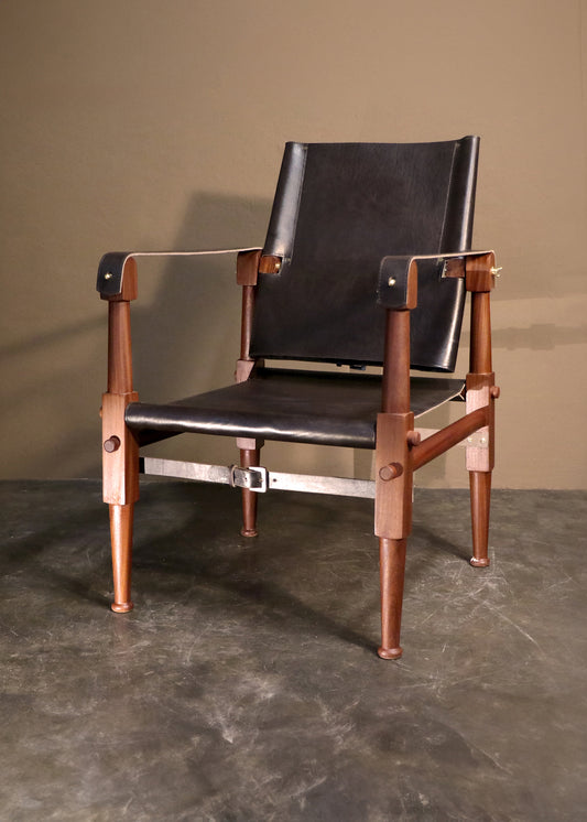 Sophisticated campaign chair with a polished mahogany wood structure and luxurious black leather upholstery, standing on a sleek grey floor with a shadowy background that highlights its sleek design and fine craftsmanship.