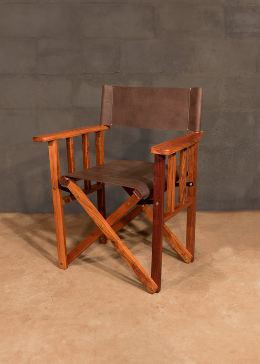 Sturdy director's chair crafted from rich African Teak with a smooth finish, complemented by a dark brown leather seat and backrest, set against a grey concrete wall, showcasing the fusion of rugged elegance and artisanal woodworking.