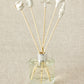 A set of white porcelain floral diffuser sticks placed in a clear glass container, with a beige background.
