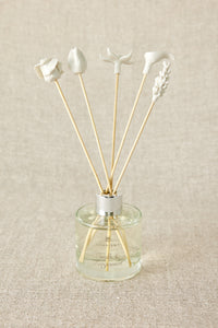 A set of white porcelain floral diffuser sticks placed in a clear glass container, with a beige background.