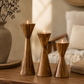 Three wooden candle holders with candles on a wooden surface, with a vase and decorative items in the background.