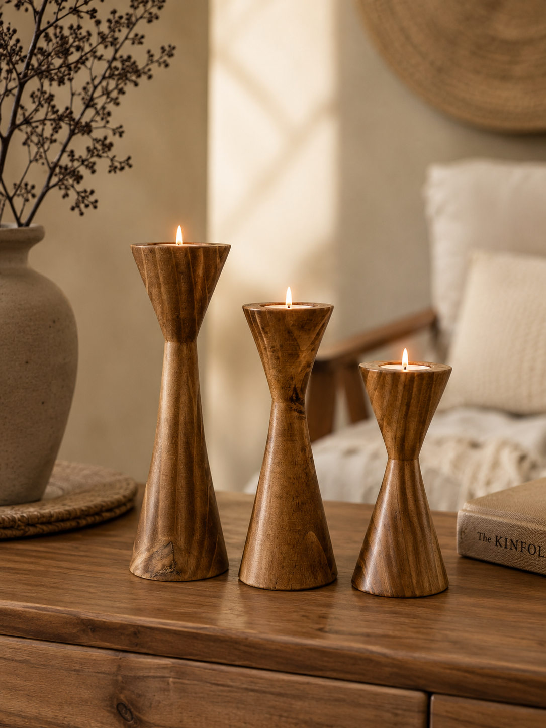 Three wooden candle holders with candles on a wooden surface, with a vase and decorative items in the background.