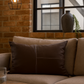 Beige sofa with brown pillow against a brick wall with a hanging light fixture.