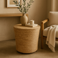 Woven side table with a plant, candle, and book in a cozy room.