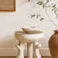 Small round table with a bowl on top, next to a vase and plant against a neutral wall.