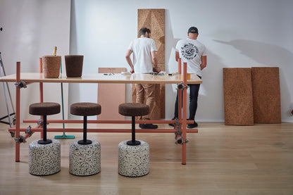 Inside the Wiid studio showroom, the Wiid Cork and Concrete Swivel Barstool in Terrazzo and Dark Cork is elegantly displayed against a backdrop of striking Wiid cork panels. This scene captures the essence of the brand's innovative design philosophy, featuring the barstool's sleek terrazzo base and comfortable dark cork seat amidst a creative installation. It exemplifies how functional art can transform any space, showcasing sustainable materials and contemporary aesthetics.