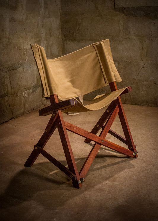 Classic Safari chair crafted with a rich mahogany wood frame and a khaki canvas seat and backrest, set in an ambient room with a textured concrete wall, exuding timeless style and handcrafted quality.