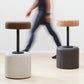 Designer Laurie Wiid van Heerden walks behind the elegantly designed Wiid Swivel Cork and Concrete Barstool in Natural Grey and Dark Cork, providing a sense of scale. This image emphasizes the stool's sleek, modern design and the contrast between the robust grey concrete base and the warm, dark cork seat. Laurie's presence highlights the thoughtful craftsmanship and sustainable innovation behind this contemporary piece of furniture.