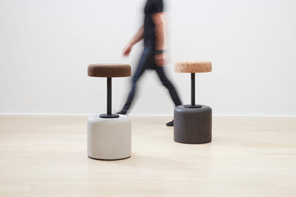 Designer Laurie Wiid van Heerden walks behind the elegantly designed Wiid Swivel Cork and Concrete Barstool in Natural Grey and Dark Cork, providing a sense of scale. This image emphasizes the stool's sleek, modern design and the contrast between the robust grey concrete base and the warm, dark cork seat. Laurie's presence highlights the thoughtful craftsmanship and sustainable innovation behind this contemporary piece of furniture.