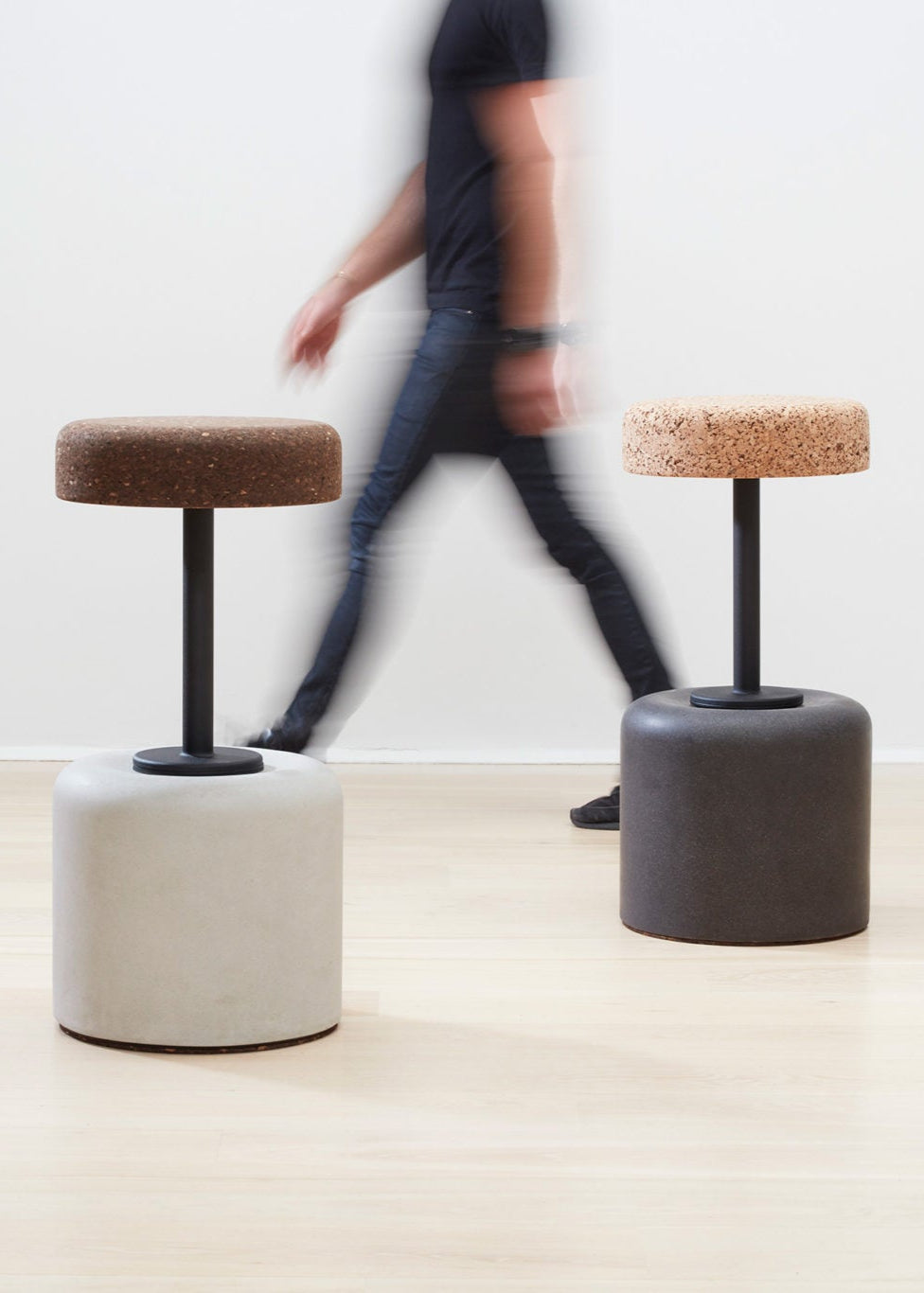 Designer Laurie Wiid van Heerden walks behind the elegantly designed Wiid Swivel Cork and Concrete Barstool in Natural Grey and Dark Cork, providing a sense of scale. This image emphasizes the stool's sleek, modern design and the contrast between the robust grey concrete base and the warm, dark cork seat. Laurie's presence highlights the thoughtful craftsmanship and sustainable innovation behind this contemporary piece of furniture.
