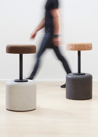 Designer Laurie Wiid van Heerden walks behind the elegantly designed Wiid Swivel Cork and Concrete Barstool in Natural Grey and Dark Cork, providing a sense of scale. This image emphasizes the stool's sleek, modern design and the contrast between the robust grey concrete base and the warm, dark cork seat. Laurie's presence highlights the thoughtful craftsmanship and sustainable innovation behind this contemporary piece of furniture.