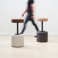 Designer Laurie Wiid van Heerden passes behind the elegantly designed Wiid Swivel Cork and Concrete Barstool in Natural Grey and Dark Cork, providing a sense of scale. This image emphasizes the stool's sleek, modern design and the contrast between the robust grey concrete base and the warm, dark cork seat. Laurie's presence highlights the thoughtful craftsmanship and sustainable innovation behind this contemporary piece of furniture