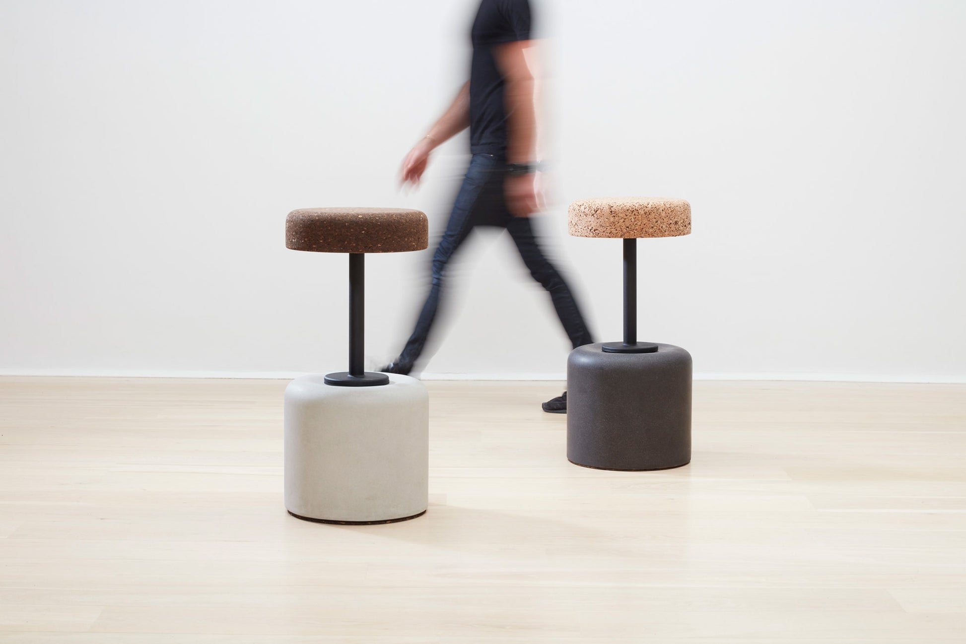 Designer Laurie Wiid van Heerden passes behind the elegantly designed Wiid Swivel Cork and Concrete Barstool in Natural Grey and Dark Cork, providing a sense of scale. This image emphasizes the stool's sleek, modern design and the contrast between the robust grey concrete base and the warm, dark cork seat. Laurie's presence highlights the thoughtful craftsmanship and sustainable innovation behind this contemporary piece of furniture